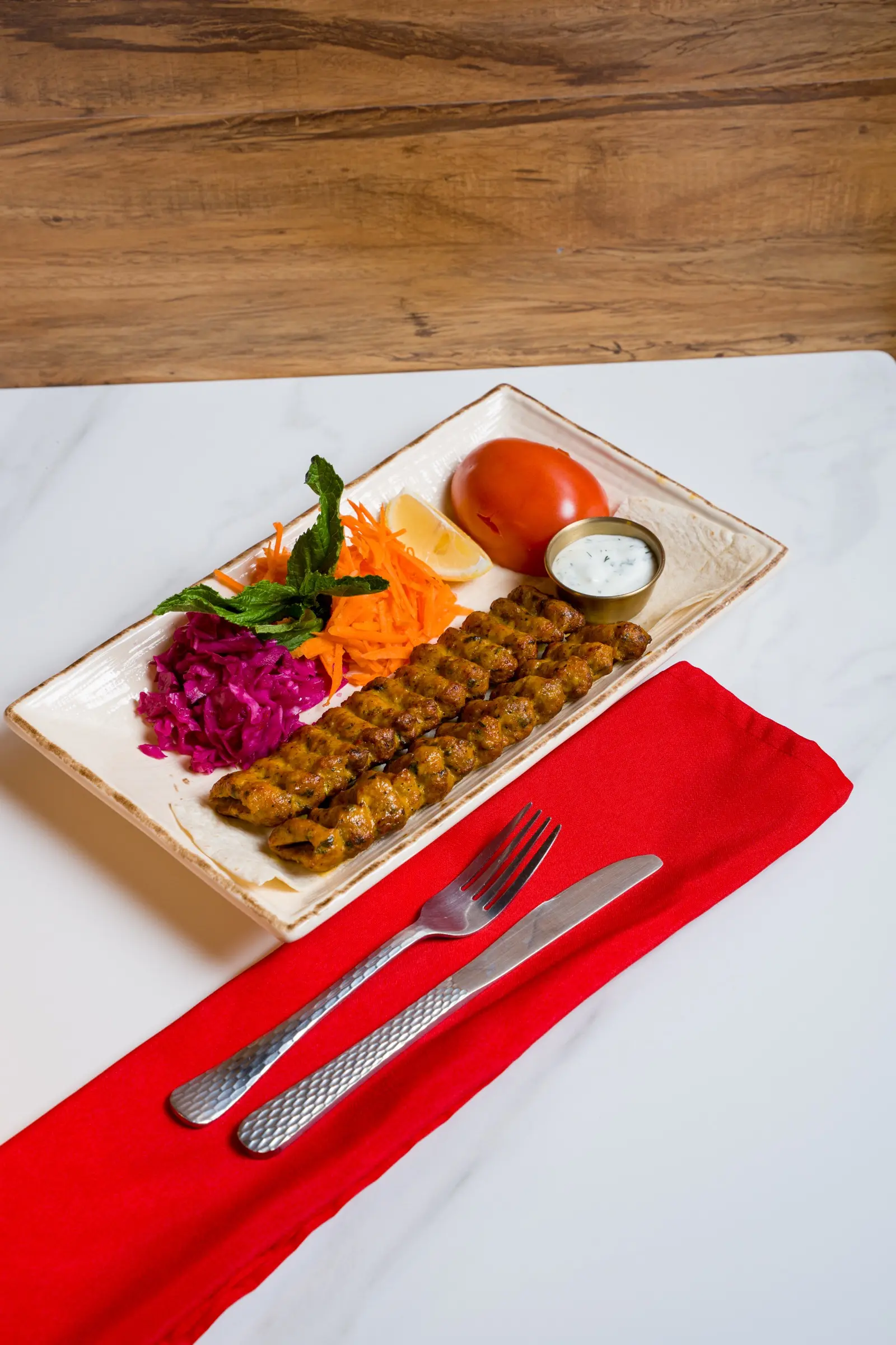 Two skewers of char-grilled seasoned ground beef with shredded carrot, red cabbage, tomato, and yogurt sauce