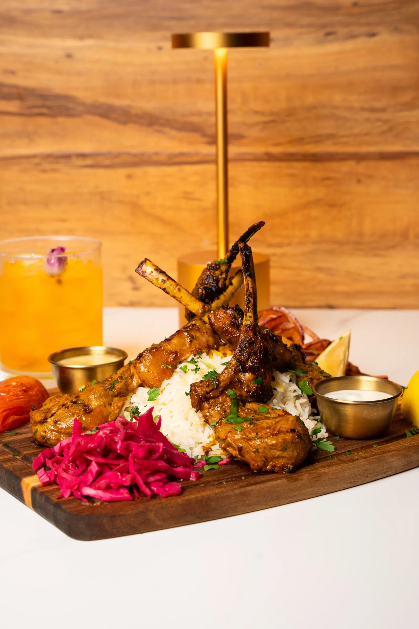 Grilled lamb chops on a bed of rice on a wooden board with pickled cabbage, sauces, and a cocktail